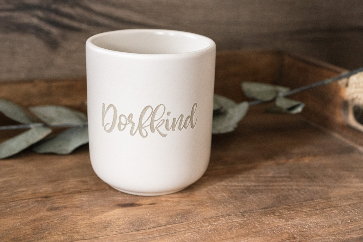 Matte weiße Keramiktasse mit Gravur „Dorfkind“ in Handlettering-Schrift auf Holzuntergrund, Eukalyptus im Hintergrund.